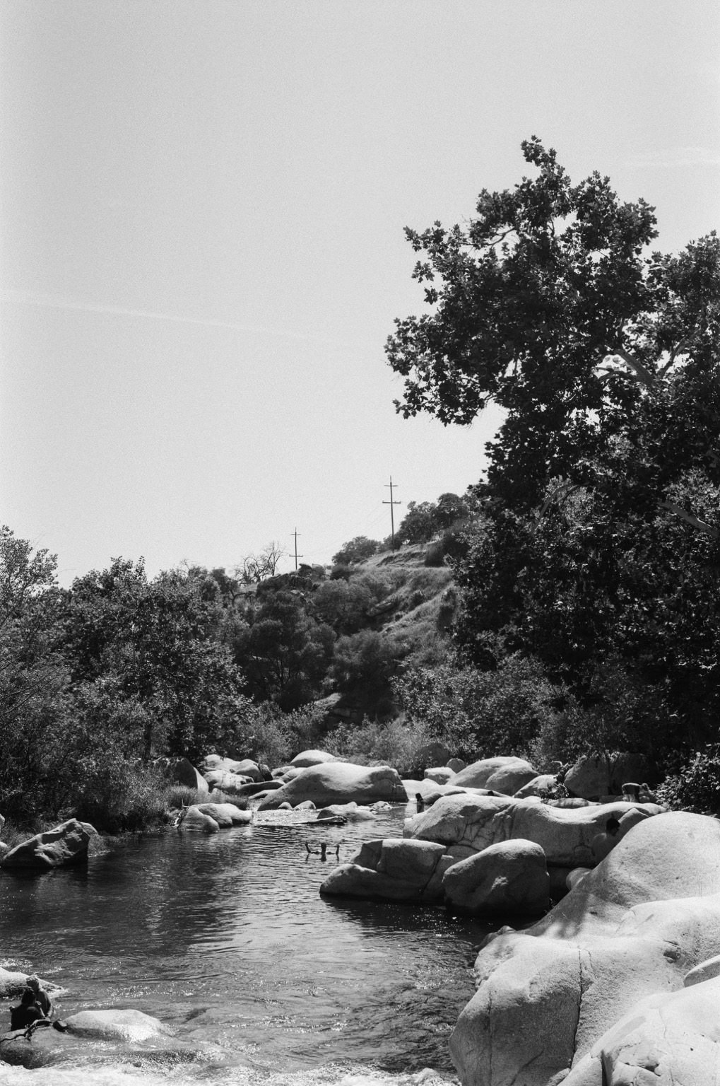 black and white 35mm film photograph of a natural landscape: a river is calm in the foreground, people made small by the scale of the scene play in the water. large rocks reflect the sun and hold shadows. trees come in from the shore. a hillside rises in the background with two poles for electrical lines rising in the distance. a bright clear sky.